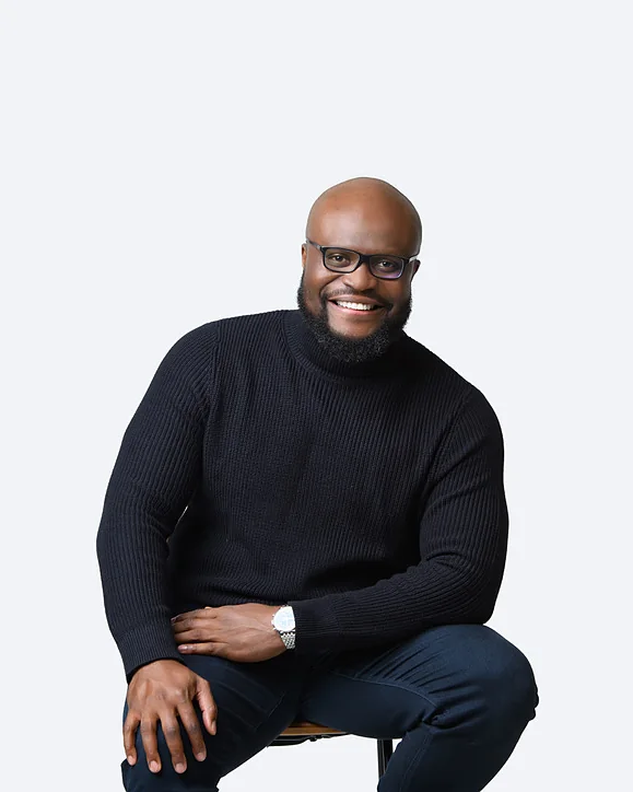 Omi, the founder, smiling, seated on a stool in a black turtleneck and glasses, one hand on his knee, against a pale studio background.